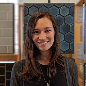 A young woman with long brown hair and a black top smiles at the camera. She stands indoors in front of a blue hexagonal tile display, with other tile samples visible in the background.