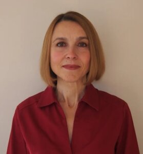 A woman with straight, shoulder-length light brown hair and fair skin smiles softly. She is wearing a collared, deep red blouse and stands against a plain beige background, looking directly at the camera.