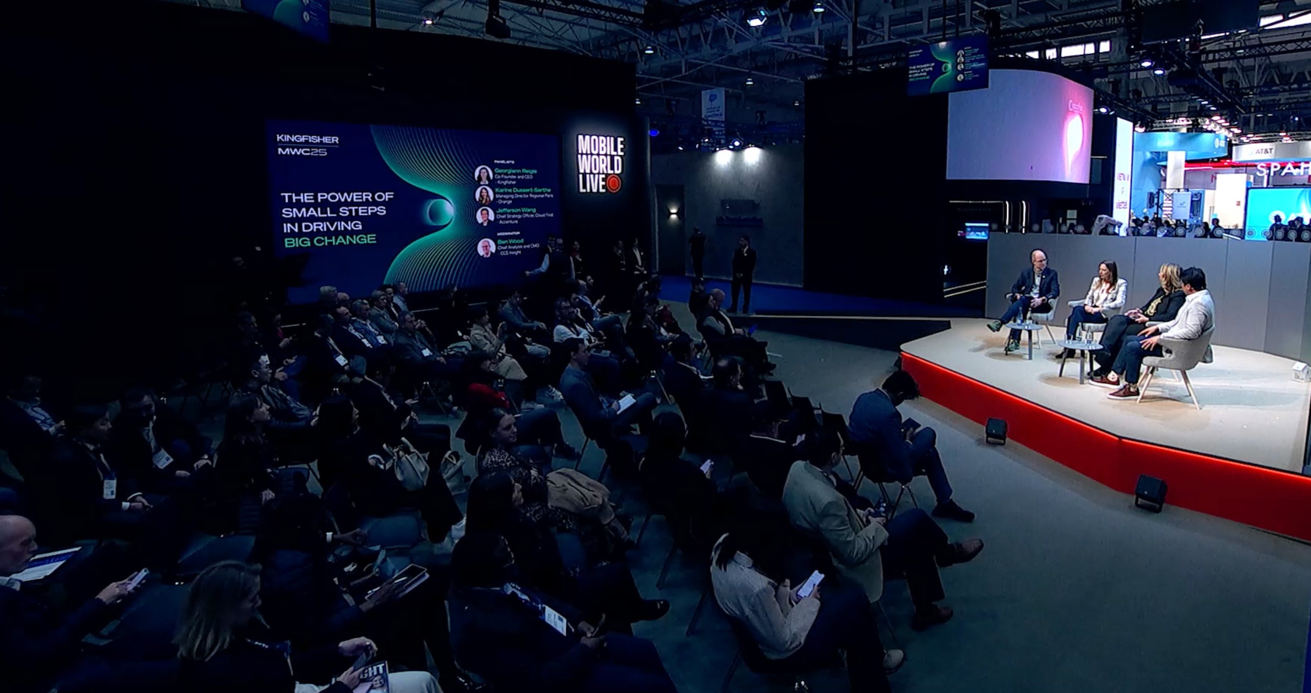 Audience seated in rows watch a panel with three speakers on a red stage. Large screen displays the topic: 