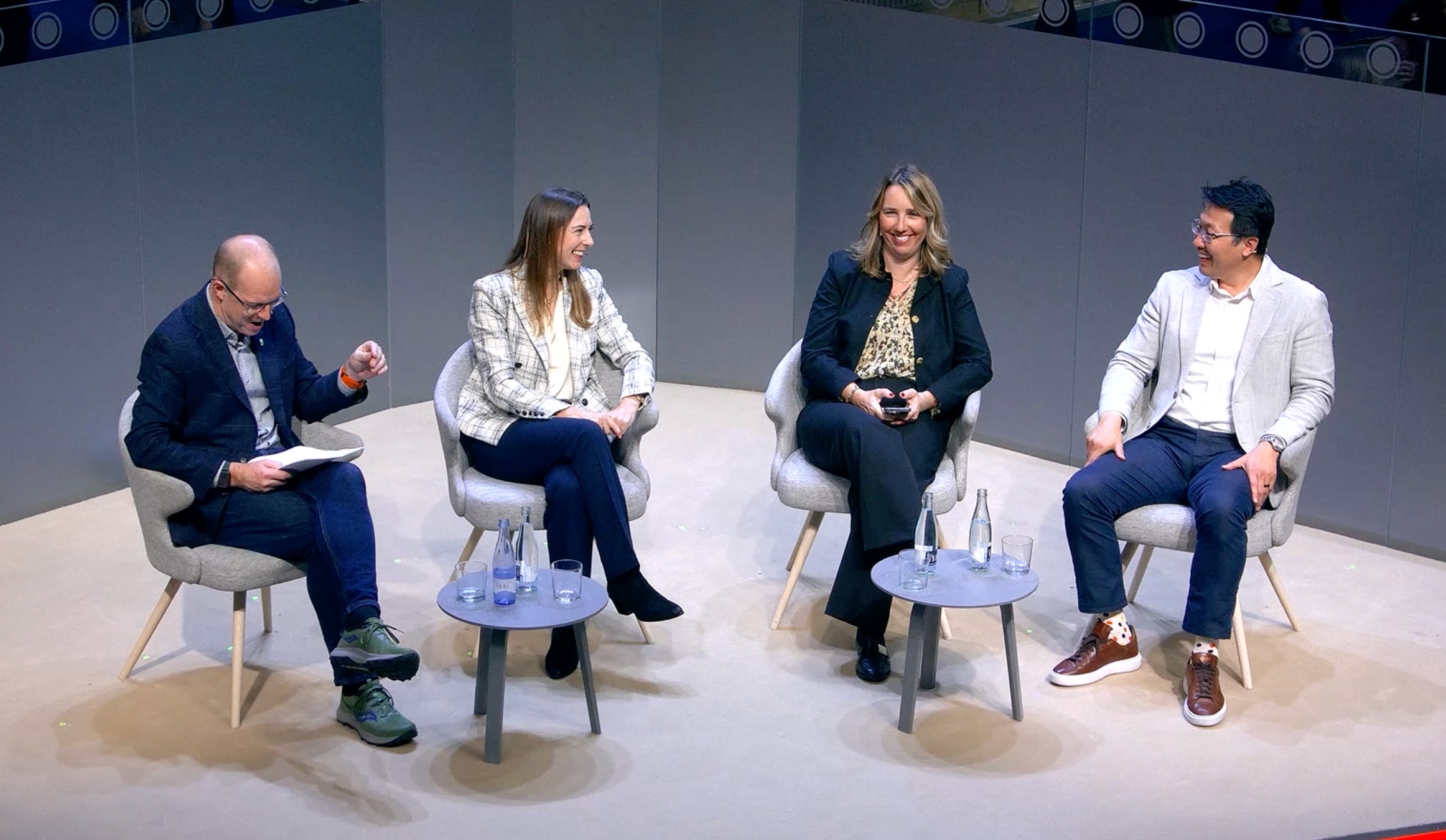 Four people are seated on stage chairs in discussion, each with a small table and water bottles beside them. The setting appears informal, with gray walls and a light-colored floor. One person is holding papers. All are engaging with smiles and attentive body language.
