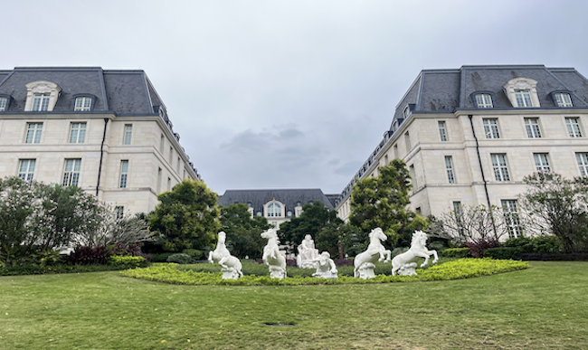 A grand, symmetrical building with slate roofs and many windows stands behind a lush green lawn. In front, white marble statues of horses and figures form a dynamic centerpiece—almost as striking as the design featured in Huawei architecture.